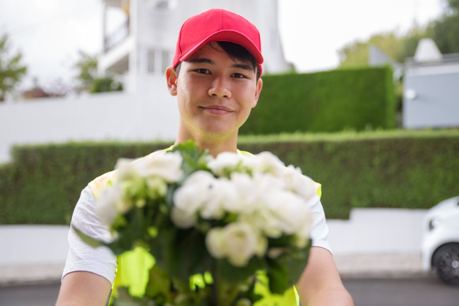 A delivery person wearing a red cap and a neon yellow vest is holding a bouquet of white flowers, including roses and lilies, wrapped in clear plastic. The bouquet appears fresh with vibrant petals an