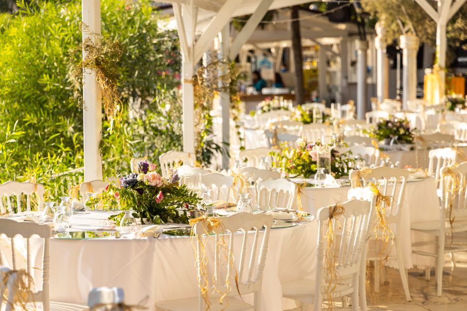 A spacious outdoor wedding reception area featuring long rectangular tables draped with white linen tablecloths, decorated with vibrant floral arrangements consisting of pink, purple, and white blooms