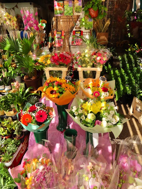 An arrangement of freshly cut flowers displayed in a flower shop, featuring vibrant bouquets of sunflowers, daisies, roses, and chrysanthemums wrapped in colorful paper and placed on tall green and wh