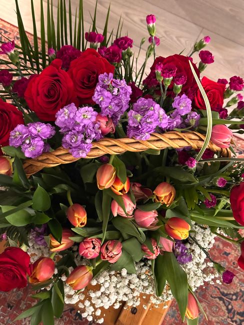 A vibrant floral arrangement featuring a mix of red roses, pink tulips, and purple statice flowers arranged in a basket with a woven handle. The bouquet showcases fresh, lush petals and sturdy green s