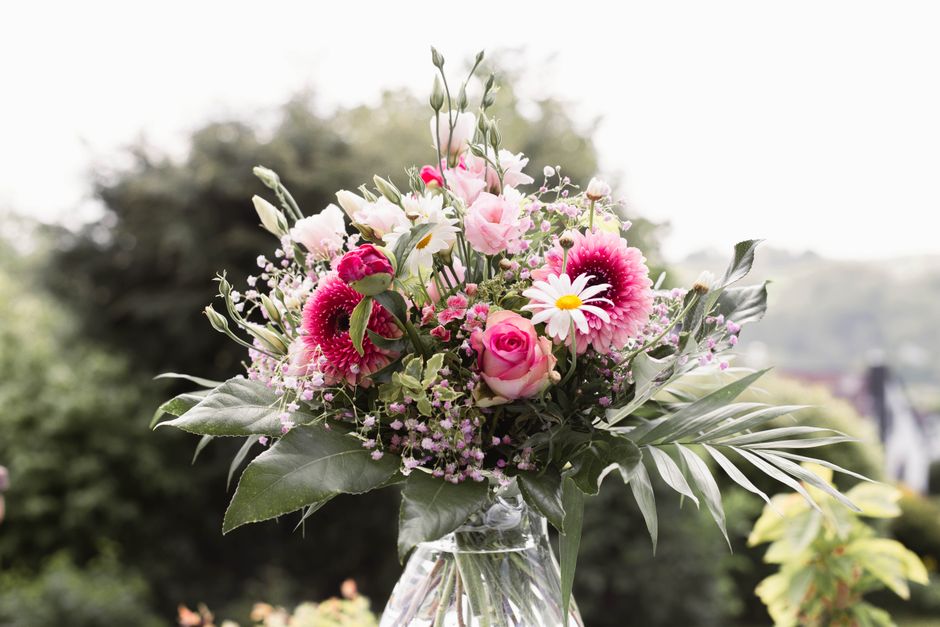 A vibrant floral bouquet arranged in a clear glass vase, featuring light pink roses, white daisies with yellow centers, deep pink gerbera daisies, and small pink filler flowers. The bouquet is accente