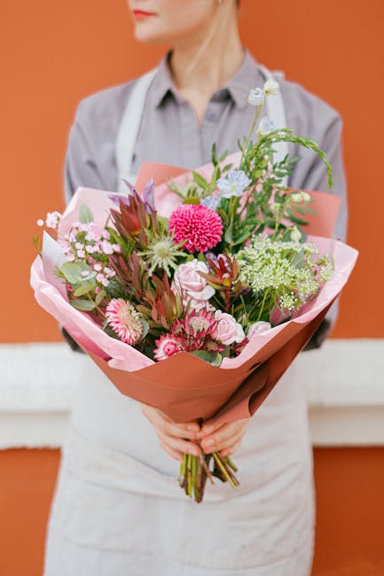 A person in a light grey shirt and white apron holds a large, vibrant bouquet of fresh flowers, including pink gerbera daisies, white chrysanthemums, pink roses, and green foliage, all wrapped in soft